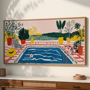 Puede incluir: Una pintura enmarcada representa una escena de piscina con una piscina azul, una silla de playa y plantas en macetas. La obra de arte presenta un cielo rosa, un patio de baldosas rojas y blancas y un sombrero amarillo. El marco es de madera marrón claro.