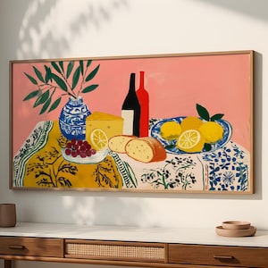 Può includere: Dipinto incorniciato raffigurante una natura morta su sfondo rosa. Una tovaglia gialla con un motivo floreale sostiene un vaso blu e bianco con foglie verdi, formaggio, uva, pane, limoni e due bottiglie di vino.