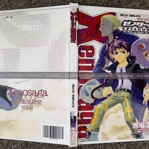 May include: A manga book cover for Xenosaga Episode I Vol 2, featuring a central character in a black jacket and green pants, a blonde character behind, and a purple-haired character. The cover includes the title, text, and a barcode.