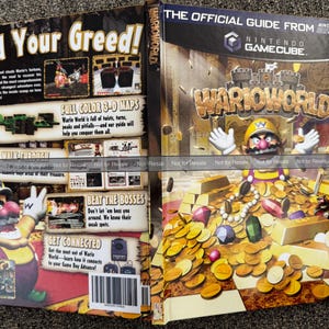 May include: A Nintendo GameCube WarioWorld game guide. The cover features Wario surrounded by gold coins and gems. The guide includes full-color 3-D maps and a complete walkthrough. The text "Feed Your Greed!" is displayed.