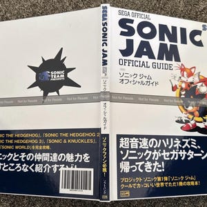 May include: A white book titled "SEGA OFFICIAL SONIC JAM OFFICIAL GUIDE" with Japanese text. The cover features Sonic the Hedgehog characters. The book is open, displaying the cover and a back cover with text and a barcode. The book is a guide for the Sonic Jam video game.