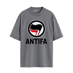 Peut inclure: T-shirt gris avec un motif circulaire : un drapeau noir avec un accent rouge, encadré par une bordure noire et blanche. En dessous, le mot "ANTIFA" est imprimé en noir. Le t-shirt a un col rond et des manches courtes.