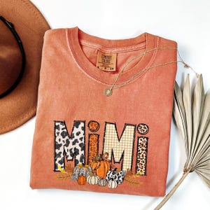 Op de afbeelding: Een koraalkleurig Comfort Colors t-shirt met het woord "MIMI" in een mix van patronen, waaronder luipaardprint en ruitjes. Het ontwerp toont pompoenen en herfstbladeren. Een gouden ketting en een bruine hoed zijn ook zichtbaar.