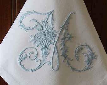 May include: A white linen tea towel with a light blue embroidered letter M surrounded by floral designs.
