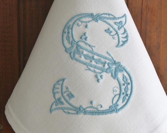 May include: A white linen napkin with a blue embroidered letter "S" in a floral design.
