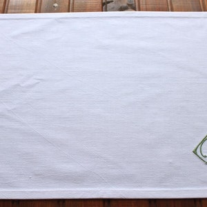 May include: White linen placemat with a green embroidered monogram "A.M." in a diamond shape.