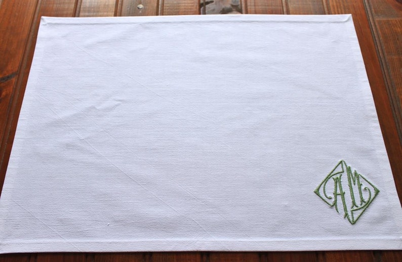May include: White linen placemat with a green embroidered monogram "A.M." in a diamond shape.