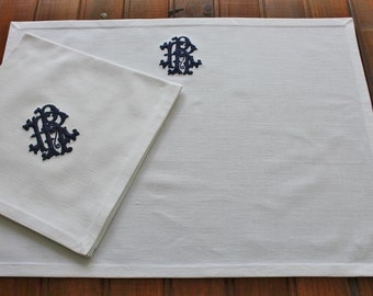 May include: White linen placemat and matching napkin set. Both pieces feature a dark blue embroidered monogram design. The placemat is rectangular, and the napkin is square. The set is displayed on a wooden surface.