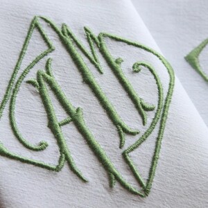 May include: A white linen napkin with a green embroidered monogram of the letters "AID" in a decorative script font.