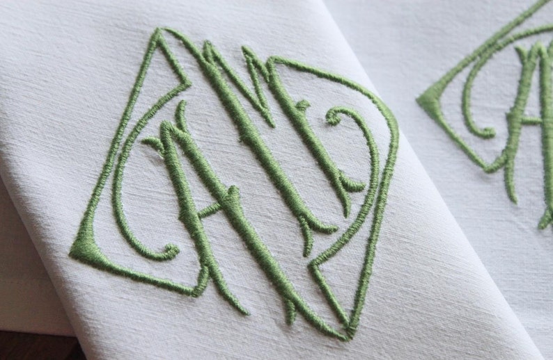 May include: A white linen napkin with a green embroidered monogram of the letters "AID" in a decorative script font.