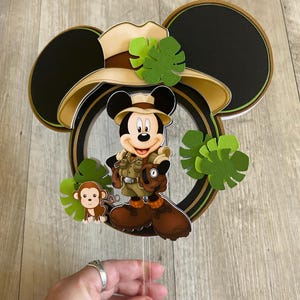 Puede incluir: Adorno para tarta con tem&aacute;tica de Mickey Mouse, con Mickey con ropa de safari, un mono y hojas verdes. El adorno tiene orejas negras de Mickey Mouse y un sombrero de safari color beige. Sobre un fondo de madera.