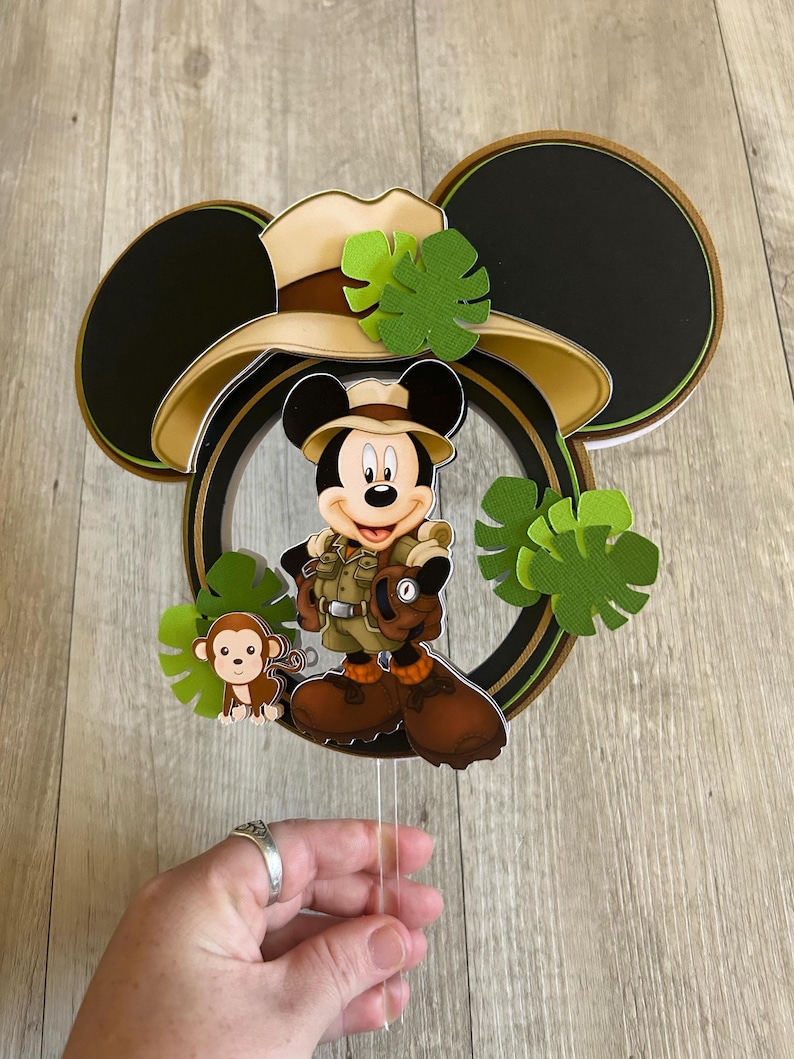 Puede incluir: Adorno para tarta con tem&aacute;tica de Mickey Mouse, con Mickey con ropa de safari, un mono y hojas verdes. El adorno tiene orejas negras de Mickey Mouse y un sombrero de safari color beige. Sobre un fondo de madera.