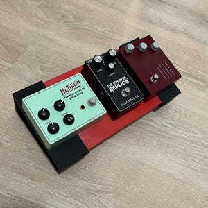 May include: A red and black guitar pedalboard with three effect pedals. The pedals are in mint green, black, and burgundy. The mint green pedal says "Benson Germanium Preamp". The black pedal says "The Schaffer Replica". The burgundy pedal has text.