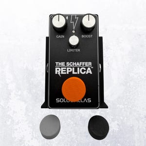 May include: Black guitar effects pedal with silver knobs labeled "GAIN" and "BOOST". The text "THE SCHAFFER REPLICA" and "SOLO DALLAS" are printed on the front. An orange circular button is centered below the text. Two replacement buttons are at the bottom.