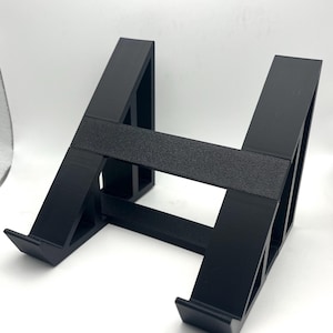 Könnte beinhalten: Ein schwarzer, H-förmiger Ständer, wahrscheinlich für ein Tablet oder ein ähnliches Gerät. Der Ständer besteht aus massivem schwarzem Material mit einem strukturierten horizontalen Balken. Das Design weist abgewinkelte Stützen und eine flache Basis auf.