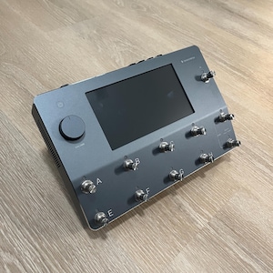 Może przedstawiać: Szare urządzenie elektroniczne z ekranem, pokrętłem głośności i kilkoma srebrnymi przełącznikami dźwigienkowymi. Urządzenie ma napis "VOLUME" i litery od A do G. Leży na drewnianej powierzchni.