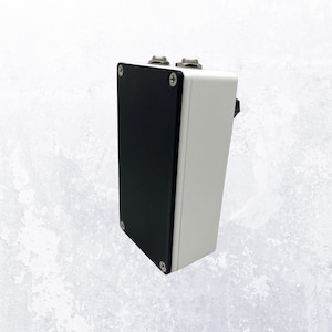 Può includere: Un dispositivo elettronico rettangolare bianco e nero con due manopole argentate sulla parte superiore. Il dispositivo ha un pannello nero su un lato ed è fissato con viti. È probabilmente un pedale per effetti per chitarra o apparecchiature audio simili.
