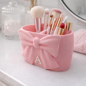Könnte beinhalten: Ein rosa Make-up-Pinselhalter mit Schleifendesign und dem Buchstaben "A". Der Halter enthält verschiedene Make-up-Pinsel, Lippenstifte und andere Schönheitsprodukte. Der Halter steht auf einer weißen Oberfläche.