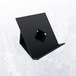 May include: A black, angled tablet stand with a diamond-shaped cutout. The stand is made of a solid material and has a small lip at the bottom to hold a tablet or phone. The stand is designed to hold a device at an angle for easy viewing.