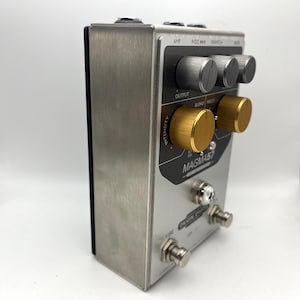 May include: A silver and black Origin Effects Magma57 guitar effects pedal. The pedal has several knobs in silver and gold, with the text "MAGMA57" and "ORIGIN EFFECTS" visible. It also features foot switches and input/output jacks.