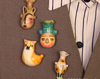 May include: A collection of four ceramic pins. The pins include a vase with a face, a dog, and two other vase designs. The pins are colorful with details like sunflowers and faces. The pins are on a gray background.