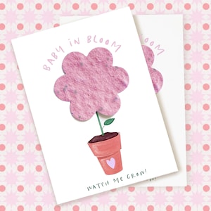 May include: Two white cards with a pink flower design. The flower is made of textured pink material and sits above a terracotta pot with a purple heart. The words "BABY IN BLOOM" and "WATCH ME GROW!" are written on the cards.