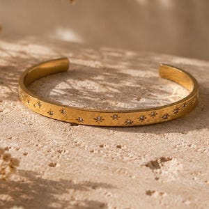May include: A gold-toned metal cuff bracelet featuring a series of engraved star motifs along the outer band. Each star design is centred with a small, clear, sparkling stone. The bracelet has an open-ended design with a smooth, polished finish.