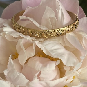 Puede incluir: Una pulsera dorada con bisagras con diseños celestiales, que incluyen soles, lunas y estrellas, está adornada con pequeños acentos brillantes. La pulsera se encuentra sobre un fondo de pétalos de flores rosas y blancas.