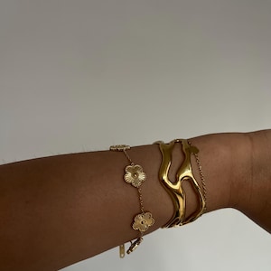 May include: Gold-toned jewelry set featuring a floral chain bracelet and a sculptural cuff bracelet. The bracelets are worn on an arm, showcasing their design and shine. The floral bracelet has three flower charms.