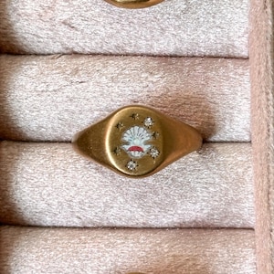 May include: Gold-toned signet ring with a seashell design. The ring features a central seashell motif in white and red, surrounded by small star and diamond accents. The ring is displayed in a pink velvet jewelry box.