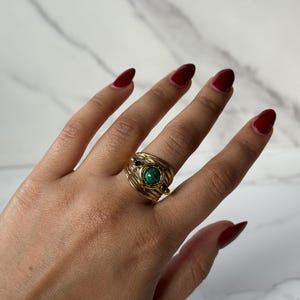 Può includere: Un anello dorato con una pietra verde centrale e pietre nere più piccole. L'anello presenta un design a filo avvolto. L'anello è su una mano con smalto rosso.