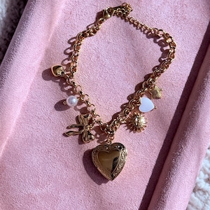 May include: Gold charm bracelet featuring a heart locket, bow, pearl, and sun charms. The bracelet is presented on a pink surface, highlighting its delicate design and intricate details. A charming piece of jewellery.