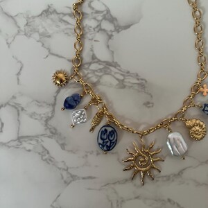 May include: Gold-toned necklace with various charms, including a sunburst, blue and white porcelain pendants, and irregular white pearls. The necklace has a chain link design and is displayed on a marble surface.