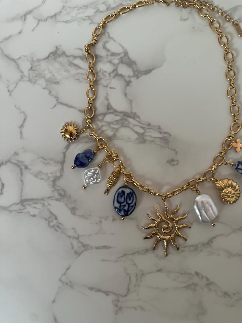 May include: Gold-toned necklace with various charms, including a sunburst, blue and white porcelain pendants, and irregular white pearls. The necklace has a chain link design and is displayed on a marble surface.