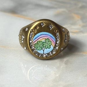 May include: Gold-coloured ring with a horseshoe design, decorated with clear crystals. The ring features a central image of a rainbow, shamrocks, and a blue sky. The ring is a signet style.