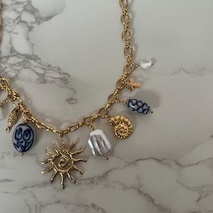 May include: Gold-toned charm necklace featuring a variety of pendants, including a sunburst, a seashell, and blue and white floral beads. The necklace has a delicate chain and a lobster clasp closure.