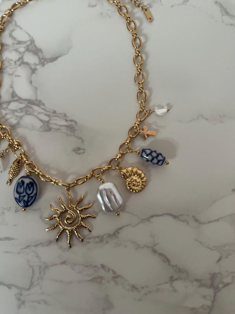 May include: Gold-toned charm necklace featuring a variety of pendants, including a sunburst, a seashell, and blue and white floral beads. The necklace has a delicate chain and a lobster clasp closure.