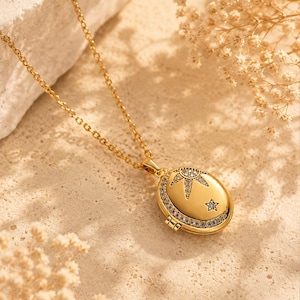 May include: A gold-toned oval locket necklace featuring a delicate chain and a pendant adorned with a sun and star motif. The locket is embellished with small, clear pavé-set crystals along the outer edge and within the celestial design.