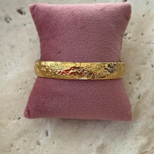 Puede incluir: Un brazalete rígido de tono dorado pulido con un intrincado patrón floral y de enredaderas en relieve que envuelve toda la superficie exterior. El metal tiene un acabado reflectante de alto brillo que resalta los grabados detallados.