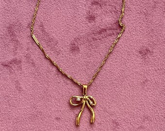 Puede incluir: Un collar dorado con un colgante en forma de lazo. El colgante tiene dos pequeñas piedras preciosas transparentes y está suspendido de una delicada cadena. El collar se muestra sobre una superficie rosa.