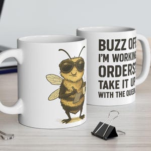 May include: Two white ceramic mugs. One mug features a cartoon bee wearing sunglasses and holding a clipboard. The other mug has the text "BUZZ OFF I'M WORKING ORDERS TAKE IT UP WITH THE QUEEN" in black. A black binder clip is in the foreground.