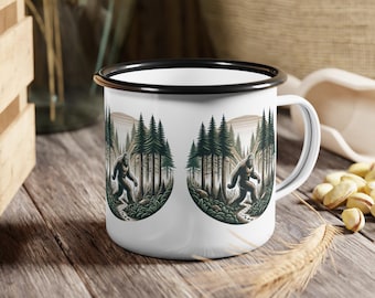 Puede incluir: Taza de esmalte blanco con borde y asa negros. La taza presenta una ilustración de Bigfoot caminando por un bosque con altos pinos y montañas al fondo. La taza está sobre una superficie de madera.