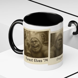 May include: White ceramic mug with a black interior and handle. The mug features sepia-toned images of a Bigfoot-like creature and forest elves. Text on the mug reads "Forest Elves '74" and "Oregon".