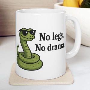 May include: White ceramic mug with a cartoon green snake wearing sunglasses and the text "No legs. No drama". The mug sits on a beige coaster. The mug is a standard size.