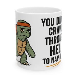 Op de afbeelding: Witte keramische mok met een cartoon schildpad met een oranje hoofdband en een humeurige uitdrukking. De tekst op de mok luidt: "YOU DID CRAWL THROUGH HELL TO NAP".