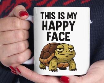 Może przedstawiać: Biały ceramiczny kubek z czarnym napisem "THIS IS MY HAPPY FACE". Poniżej znajduje się rysunkowy żółw z brązowym pancerzem i uśmiechniętą twarzą. Kubek jest trzymany przez osobę.