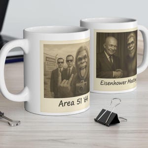 May include: Two white ceramic mugs with images printed on them. The mug on the left has a black and white photo of three men and a large-footed creature with a camera. The text "Area 51 '64" is printed below the image. The mug on the right has a black and white photo of a man in a suit and a large-footed creature. The text "Eisenhower Meeting" is printed below the image.