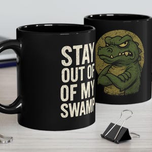 May include: Two black ceramic mugs. One mug features the text "STAY OUT OF MY SWAMP" in white. The other mug has a green alligator illustration with crossed arms against a yellow circle.