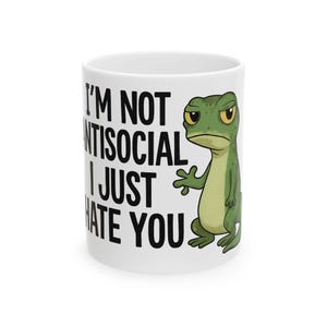 May include: White ceramic mug featuring the text "I'M NOT ANTISOCIAL I JUST HATE YOU" in black. A cartoon gecko in green and yellow is on the right. The gecko has a grumpy expression.
