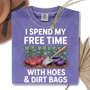 a purple t-shirt with a graphic design depicting a garden scene, including tomatoes, eggplants, and a shovel. The text on the t-shirt reads "I SPEND MY FREE TIME WITH Hoes & Dirt Bags".